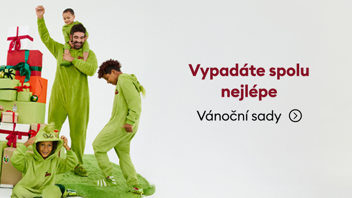 Skupina lidí v zelených vánočních kostýmech stojí vedle barevných dárků. Text na obrázku říká: 